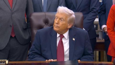 Donald Trump listens to a reporter asking a question from behind his desk in the Oval Office.