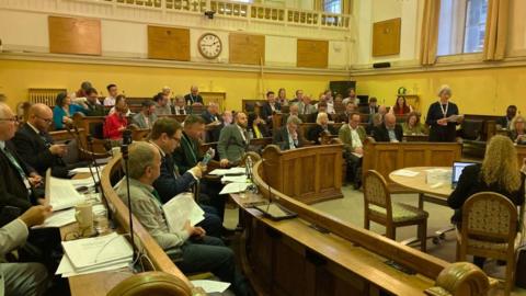 A council meeting is taking place. A woman is standing and speaking.