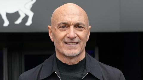 Simon Milton smiles at the camera inside Portman Road stadium. He is bald and has a shaved beard. He wears a black jacket with a black top underneath.