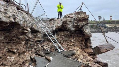 A man in a high-viz anorak and hard hat is standing at the top of the pier section which has collapsed. Metal fencing has fallen down and there are rocks and boulders strewn everywhere.