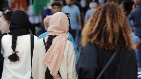 Three Iranian women, one wearing a black head covering, another with a pink hijab and a fourth one without a head covering with thick curly brown hair, walking.