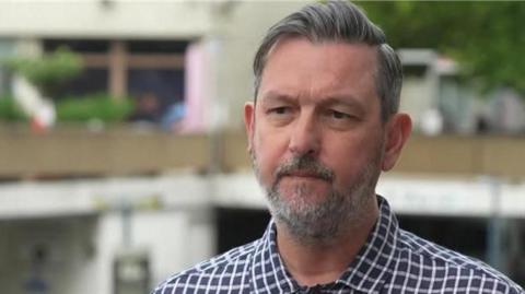 Chris has grey hair and a grey beard. He is looking just off camera and is wearing a chequered blue and white shirt.