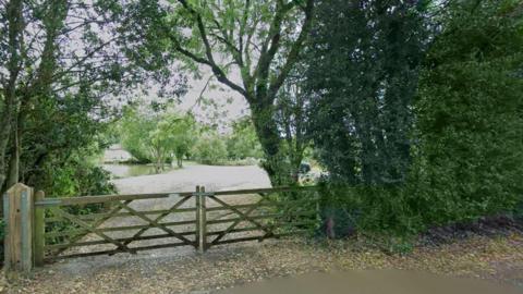 A pond and nature area seen from a road. A wooden double gate bars the entrance. Trees and bushes can be seen lining the site and by the pond. Leaves are strewn on the ground.