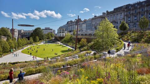 A view of Union Terrace Gardens showing the plant arrangements, a grassy area in the centre and people walking around or sitting on the grass.