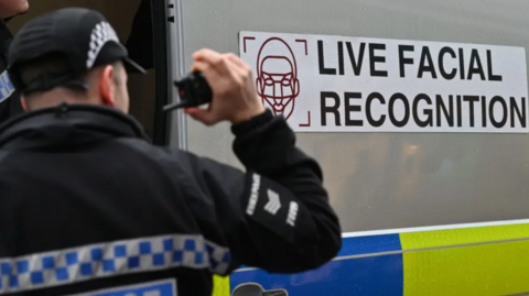 A police officer holds a radio while standing next to a van which bears the words "Live Facial Recognition"