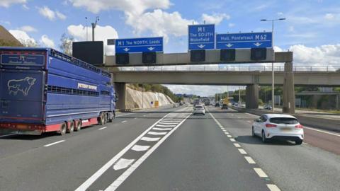 A motorway junction with five lanes marked out. A large blue livestock lorry in the far left lane. Two white cars in the middle lanes. A gantry over the road with three blue signs. One says M1 North Leeds, the next says M1 The South, Wakefield. The third says Hull, Pontefract M62.