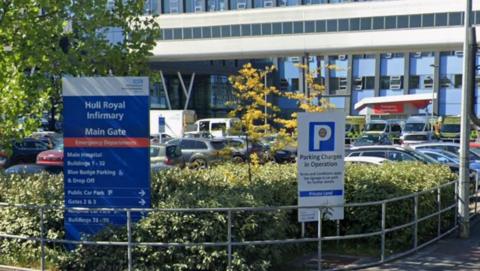 The entrance to Hull Royal Infirmary car park. It shows a sign reading "Hull Royal Infirmary Main Gate" with a list of buildings below. On the right is a parking sign. The signs are nestled in bushes and cars can be seen parked behind the signs, in front of the hospital building.