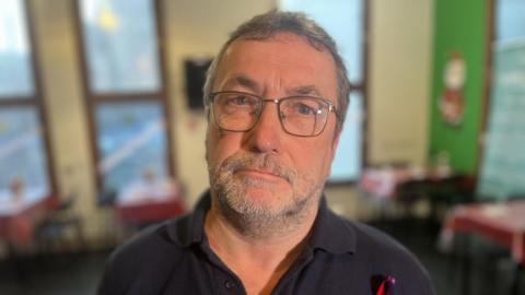 Damian, who has short, tousled grey hair and stubble, wears silver square-rimmed glasses. He is wearing a polo-shirt open at the neck and looking unaffectedly into the camera.