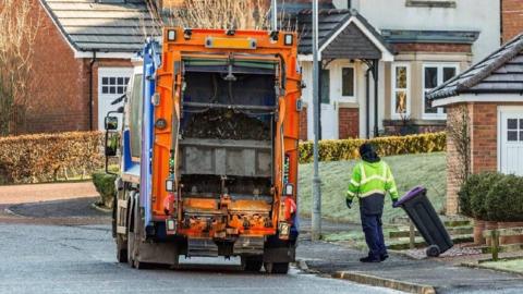 An partially-orange coloured bin lorry, with a council worker in hi-viz stood to the right, holding a wheelie bin at an angle.