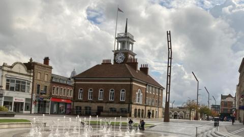 Stockton Town Hall. It is a stone and brick building with windows running alongside the front and sides. It has a clock positioned on its tower and a flag flying from a pole. The area in front has a display of fountains, while shops can also be seen along High Street.