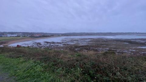 Coastline at Vazon in Guernsey. It's a murky day with brown coastal vegetation and sand in the distance. Grey skies with white cloud