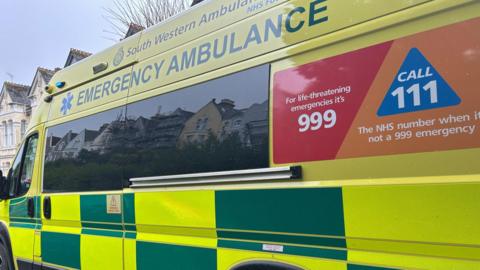 A side view of an ambulance belonging to the South Western Ambulance Service. It is bright yellow and green with a red and orange poster reading 'for life-threatening emergencies it's 999, call 111 the NHS number when it's not a 999 emergency. A few three-storey houses can be seen at the front of the ambulance.