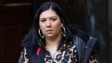 A woman with long dark hair, large earrings and a number of piercings in a leopardskin pattern blouse and dark gilet