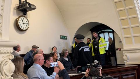 A police officer standing in front of a group of people sitting down in rows in a council chamber. Several security guards dressed in hi-vis jackets are standing under an archway by a door. One person is taking a picture of the officer with their phone while another is taking a picture with their camera.