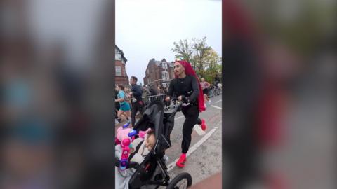 Aneila Afsar runs while pushing her daughter in a pram. She wears a black tracksuit with red headscarf and trainers