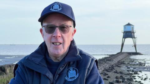 Andy Schooler is wearing glasses and a navy cap, jumper and gilet. Behind him, a rocky causeway leads to one of the lighthouses, which is mostly surrounded by water has a white building on top of iron legs.