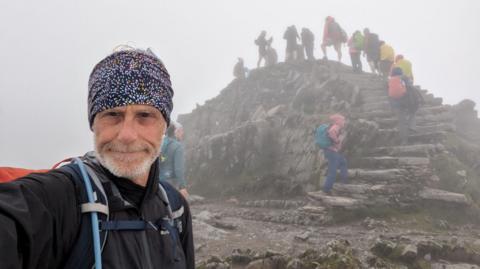 Andrew Lamb in selfie taken at top of mountain in cloudy conditions with people behind walking up to the very top on steps in mountain gear