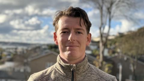 Max Andrews smiles at the camera. He is wearing a grey funnel neck fleece. He has slicked back black hair. St Helier is visible in the background, along with the coastline.