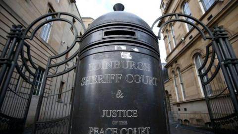 The black gates at the entrance of Edinburgh Sheriff Court. In the centre is a large bollard which has white print reading "Edinburgh Sheriff Court and Justice of the Peace Court".