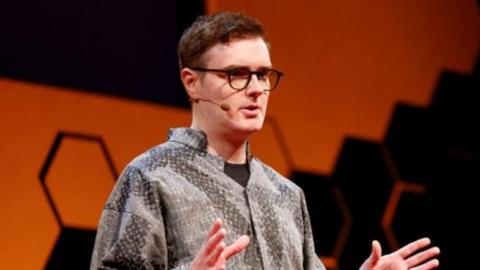 Welsh computer scientist Llion jones stands on stage at a TED-AI conference in San Francisco, wearing a grey patterned, loose-fitting shirt. The background features a bold orange shape with a honeycomb-style black geometric design. Llion wears glasses and a microphone and is mid-presentation, with both hands raised in a gesturing position.