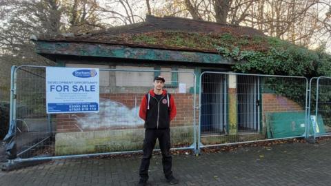 Simpa Carter is standing in front of the fenced-off toilet block. The one-story brown-brick building has a triangle roof and green door and window panes. A FOR SALE sign is attached to the fence. Simpa is wearing black jeans and black hoodie with red sleeves, and a cap.