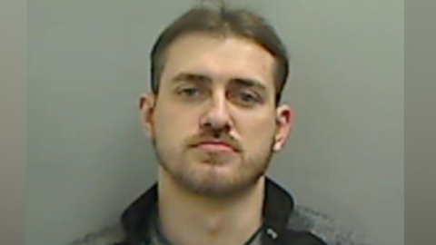 A mugshot of Dale Hamilton. He is looking into the camera with a slight frown. He has dark, short hair and a beard. He is wearing a black and grey zippy top.