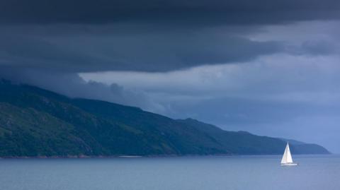 A yacht with a white sail in the Sound of Mull off the Isle of Mull under dark clouds.