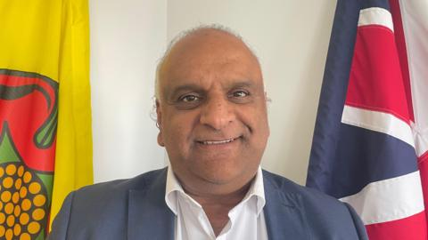 An image of County Councillor Azhar Ali, who is wearing a blue suit, with a white open necked shirt. He is smiling. He is standing in front of the union flag to his right and the Lancashire flag to his left.