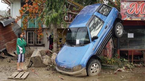 A young woman in a green and white jacket looks on forlornly as a blue car hangs from a tree, with its rear end in the air, having been lifted by floods during Typhoon Rai in the Philippines in 2021