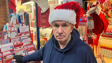 John Bird looks fed up, he is wearing a red Santa hat and a blue fleece top with a hood, he has his right hand out on a selection of Christmas cards. He is in the doorway of his shop with lots of festive cards and items visible in the background.