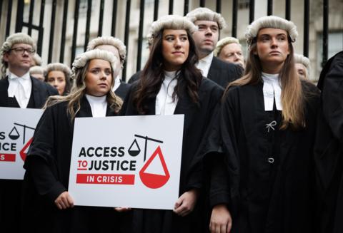 A row of barristers wearing black robes and wigs stand in front of a set of black iron railings. The three at the front are woman with long brown hair, behind them men can be seen. The woman in the middle is holding a placard which has a stylised scales of justice on it and reads "Access to Justice In Crisis"