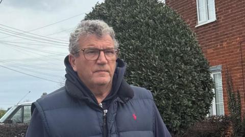 Terry Penton has grey hair and is wearing glasses, a navy bodywarmer and hoodie. He looks serious and is standing outside a brick house.