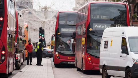 A busy central London street with multiple red double-decker buses stuck in slow-moving traffic. A white van and several cars are also queued, while two men stand on the pavement beside a bus. Christmas lights hang above the road, and buildings line the background.