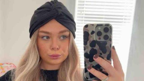 Ellie wearing a blonde wig and a black headband is taking a selfie on her iphone in the mirror.