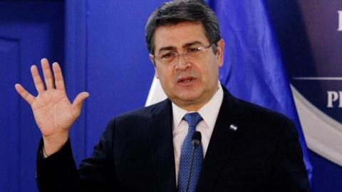Honduras' President Juan Orlando Hernandez speaks at the Presidential House in Tegucigalpa, Honduras in January 2020. He is wearing a suit and navy blue tie, behind him is a royal blue backdrop. He is gesturing with his right hand.