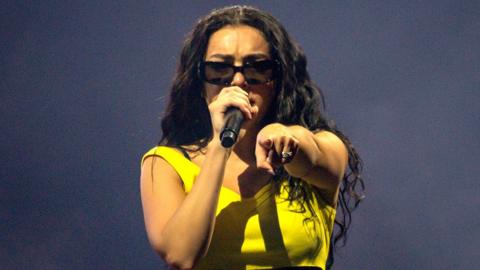 Charli XCX wears a yellow strappy top. She has long brown wavy hair and is wearing black sunglasses. She is singing in to a microphone and pointing at the camera.