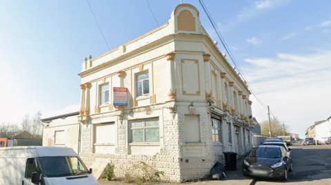 A closed pub in Bradford. The building sits on a road junction and is painted white with boarded-up windows and a for sale sign on the second storey.