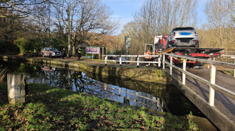 A bridge over the Leeds-Liverpool Canal near Saltaire. A truck on the bridge has a silver Range Rover on its flatbed