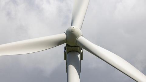 Close-up of a wind turbine’s rotor and three blades against a cloudy sky, viewed from below.