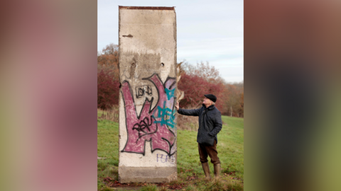 A man in a dark coat, brown trousers, wellington boots and a black cap stands next to a thin section of concrete that has pink, blue and black graffiti on it.