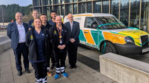 Students dressed in paramedic uniforms stand in front of an ambulance car. There are two women and five men pictured. Two of the men are in suits.