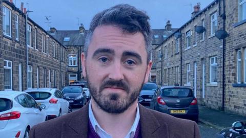 A man with a dark beard and silver/grey hair wearing a brown suit jacket and purple V-necked sweater standing in a terraced street.