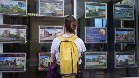 A woman stands with her back turned looking at the front window of an estate agent.