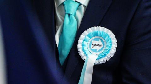 A blue and white Reform UK rosette is pinned to a dark blue suit with a light blue tie