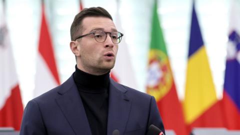 Bardella speaks while wearing a dark turtleneck and suit in front of European flags, at the European Parliament in October.