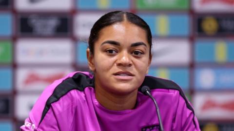 Footballer Jess Carter sits at a desk speaking into a microphone. She is wearing a purple and black jersey.