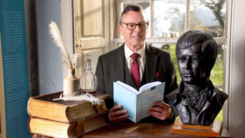 A man in glasses with a red tie and a brown jacket and cream shirt opens a book next to a bust of Robert Burns and some books and quill pens