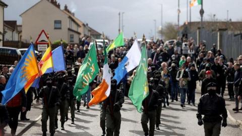 Shows masked men in paramilitary-style uniforms and carrying various flags taking part in the illegal parade in 2023