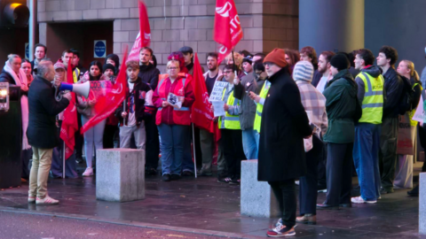A number of people gathered at a picket line. One man has a loudspeaker and other people have banners and flags.