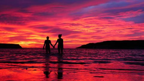 Photo of red skies above the sea with hills in the background in the shadow. In the foreground are two people, hidden in darkness, holding hands and walking into the sea. The waves are reflecting the skies.